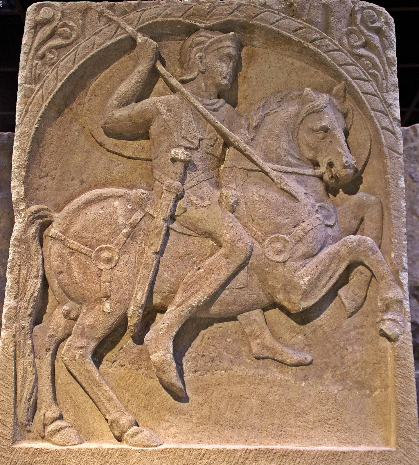 A carved stone relief of a Roman cavalryman on a rearing horse, cloak streaming, thrusting a spear downward.