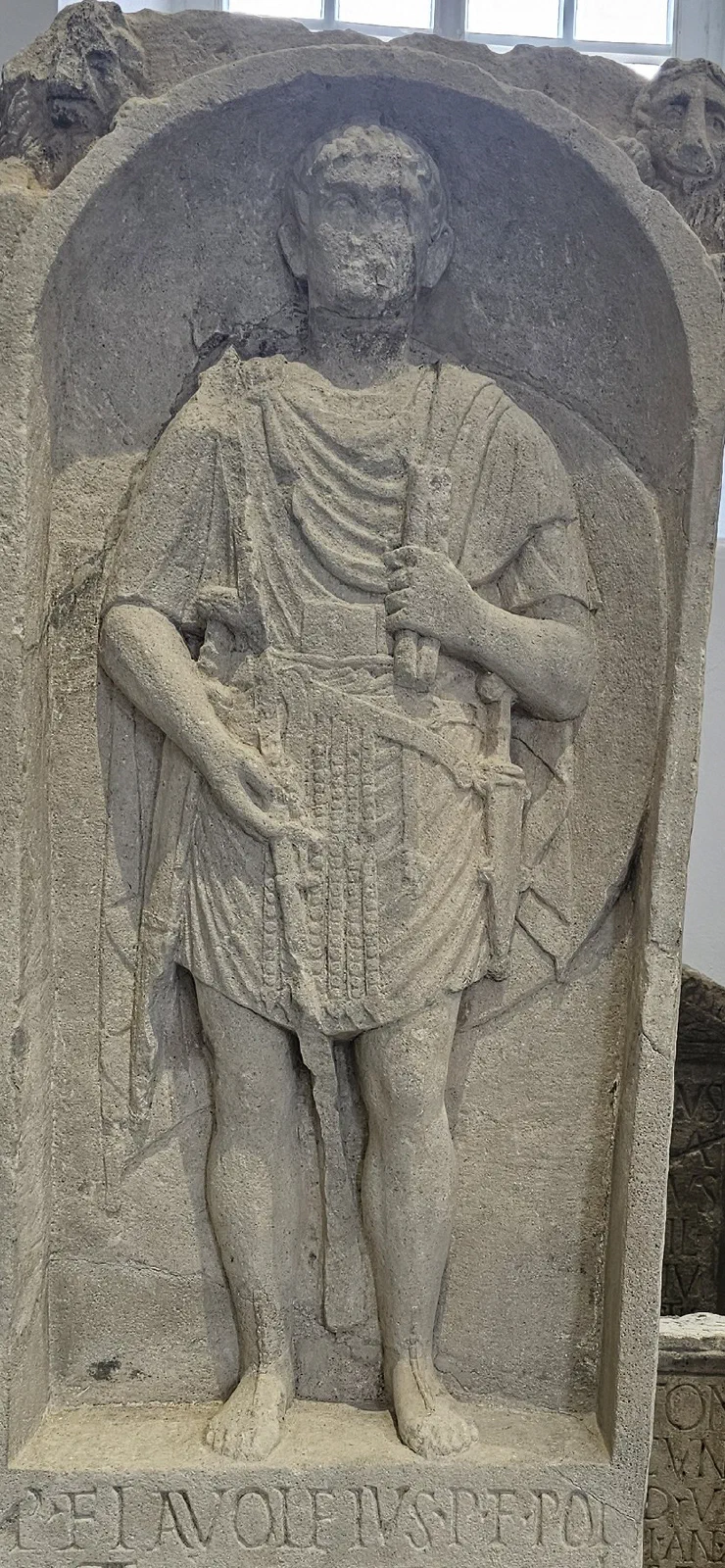 A carved stone tombstone: a Roman legionary standing in an arched niche, a large woollen cloak sweeping from one shoulder across to his other arm, a sheathed sword at his hip.